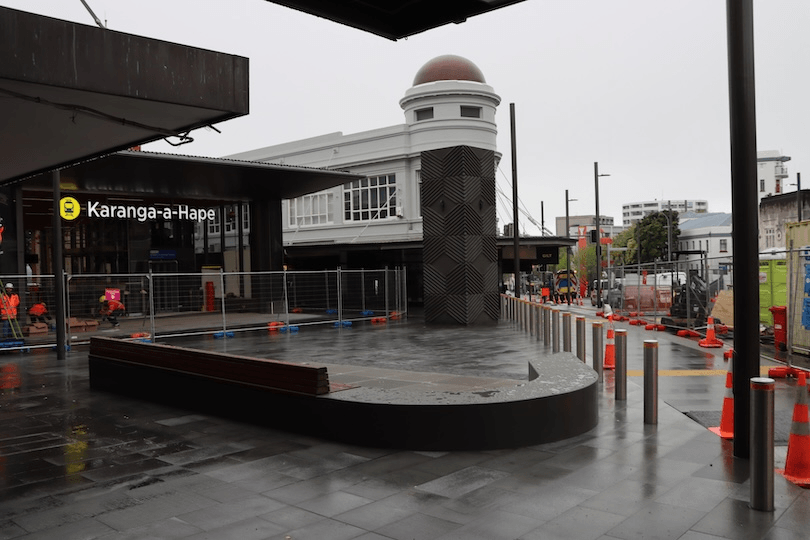 A rainy street scene shows the Karanga-a-Hape station entrance, surrounded by construction barriers and orange traffic cones. Modern and older buildings are visible, with wet pavement reflecting the overcast sky.