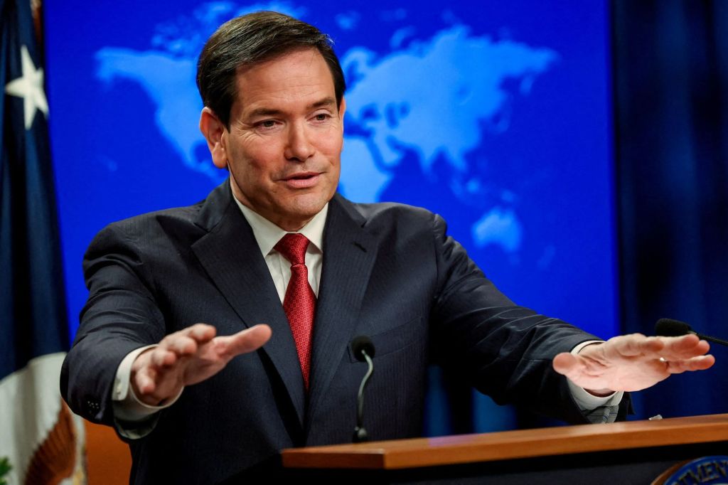 U.S. Secretary of State Marco Rubio speaks at a podium with his hands raised, in front of a blue screen with a world map and a U.S. flag