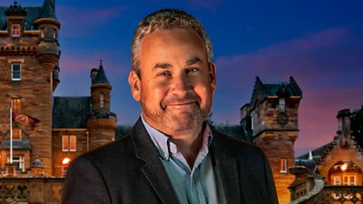 A man in a light blue shirt and dark blazer smiles to camera, set against the backdrop of a castle by night