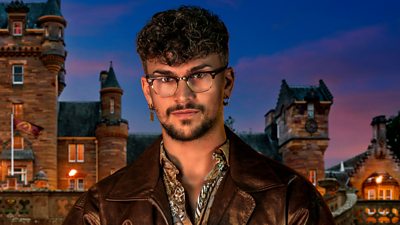 A man in a patterned shirt and leather style jacket, wearing spectacles, is set against the backdrop of a castle by night