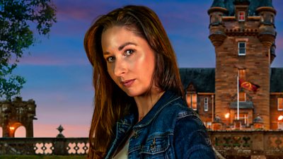 A woman with long brown hair looks to camera, side on, wearing a denim jacket and green top. She's set against the backdrop of a castle by night.