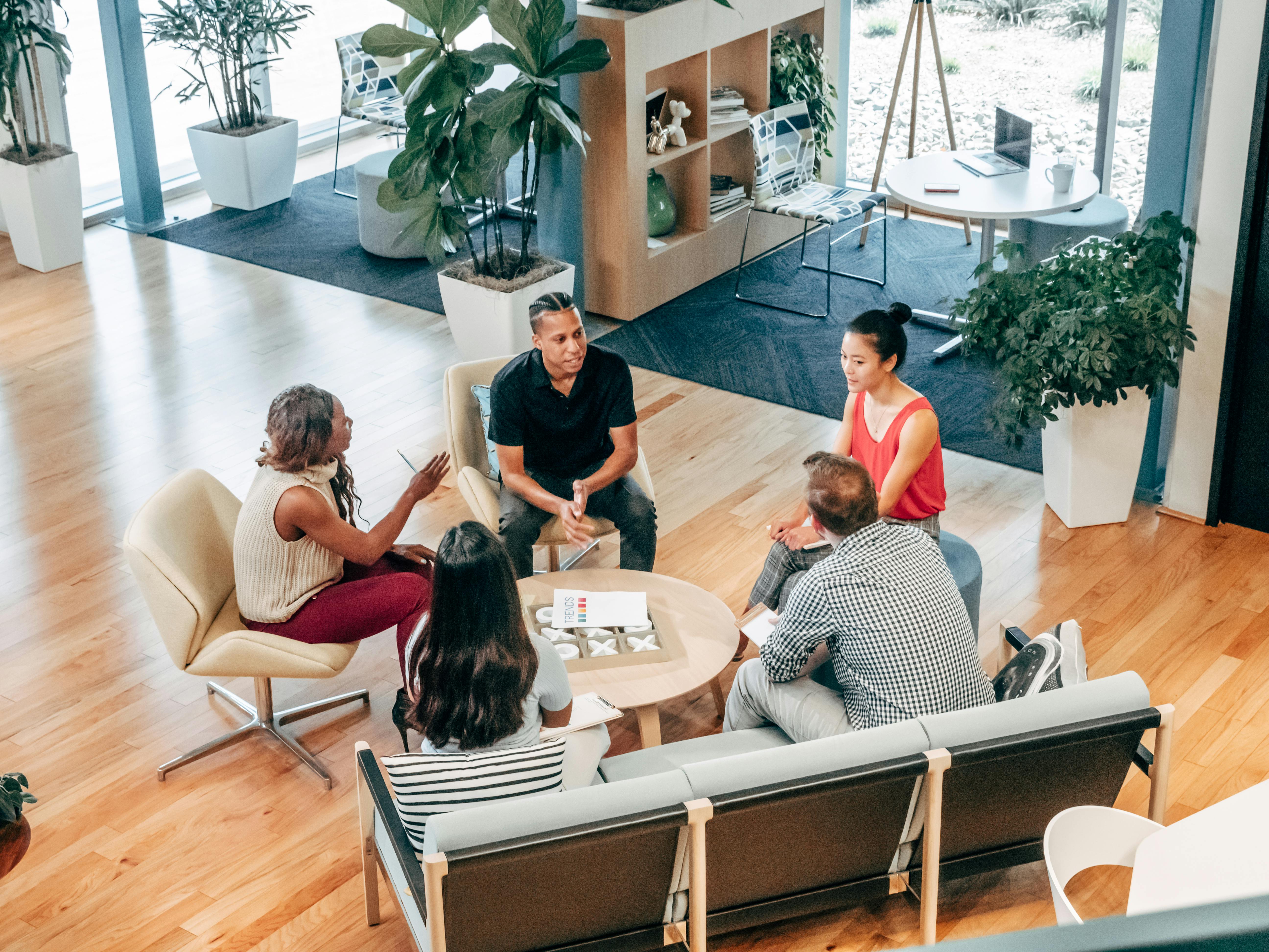 A diverse group of coworkers sit in a circle discussing ideas in a modern office lounge with plants and casual seating.