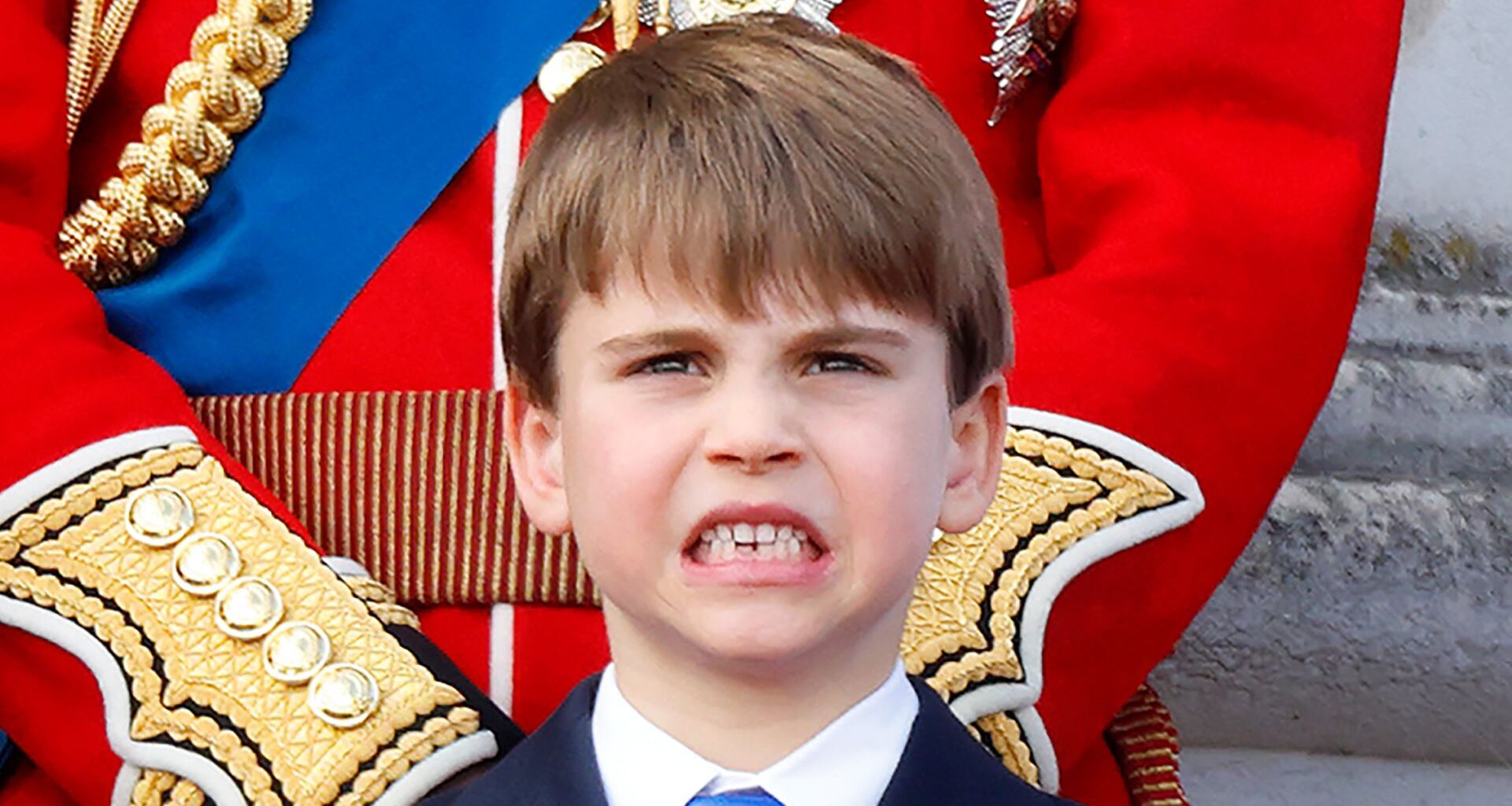 Prince Louis stands on the Buckingham Palace balcony wearing a navy suit with a white shirt and royal blue tie