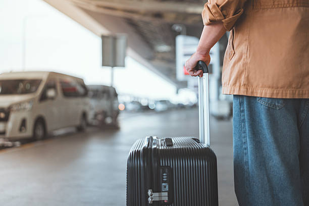 Traveler pulls a rolling suitcase along an airport curbside with vehicles parked nearby.