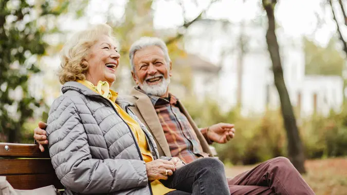 A happy senior couple sitting on a bench outside during winter weather together in warm clothing.
