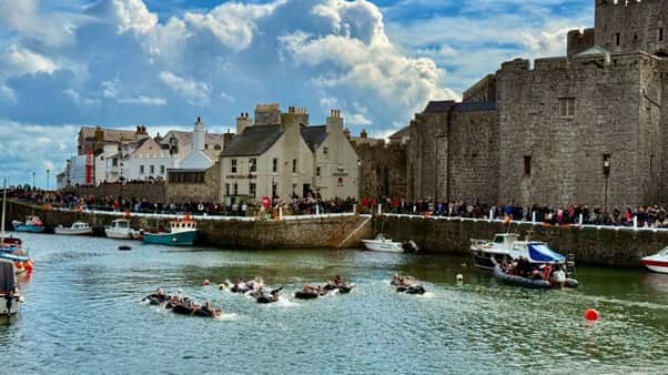 Hundreds of people crowd around old medieval castles and a waterfront at an island. People also drive boats and swim in the blue waters.