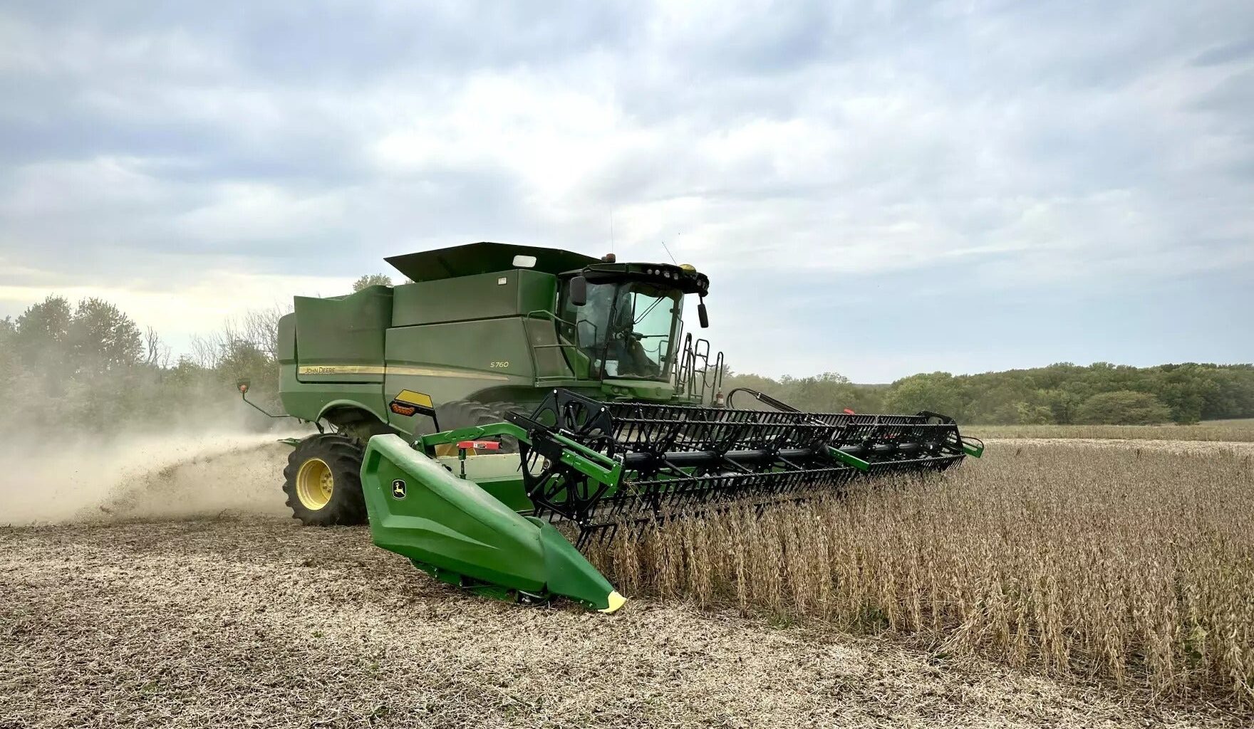 A green combine harvester is harvesting a field of crops under a cloudy sky, with dust trailing behind the machine.