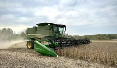A green combine harvester is harvesting a field of crops under a cloudy sky, with dust trailing behind the machine.