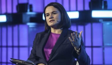 A woman wearing a dark suit and a bright purple top speaks while seated on a stage against a backlit grid.