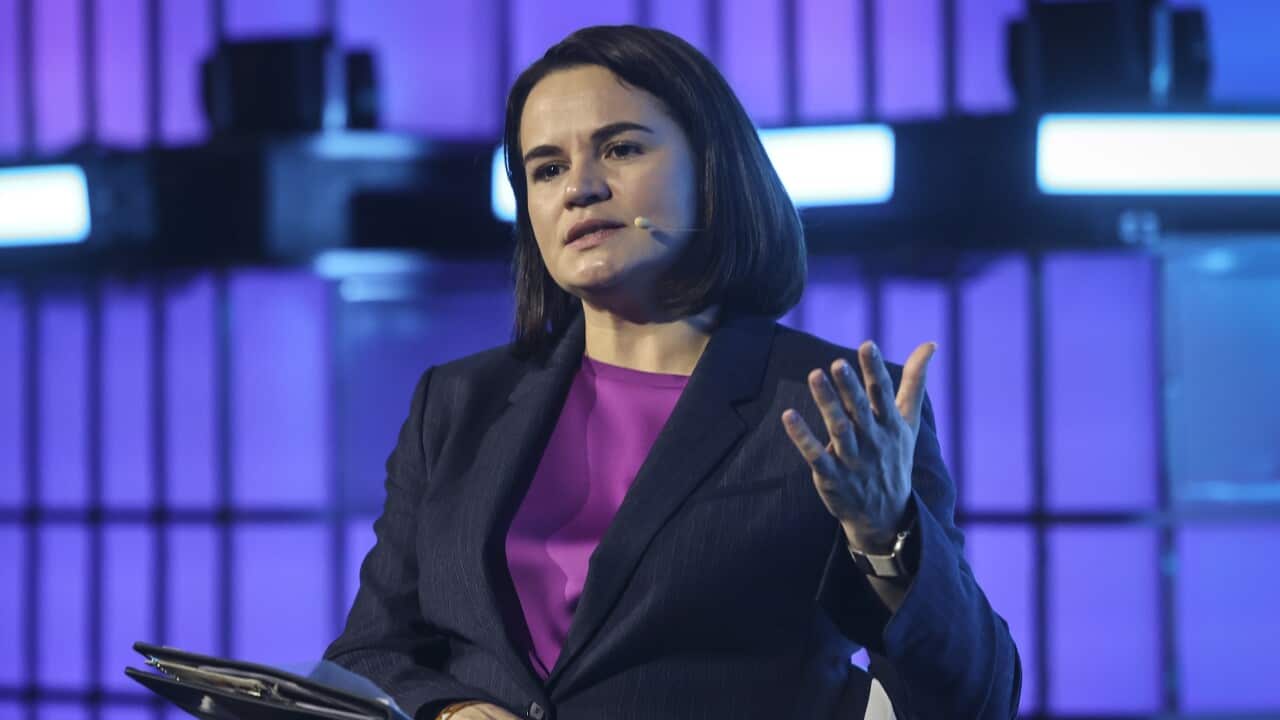 A woman wearing a dark suit and a bright purple top speaks while seated on a stage against a backlit grid.