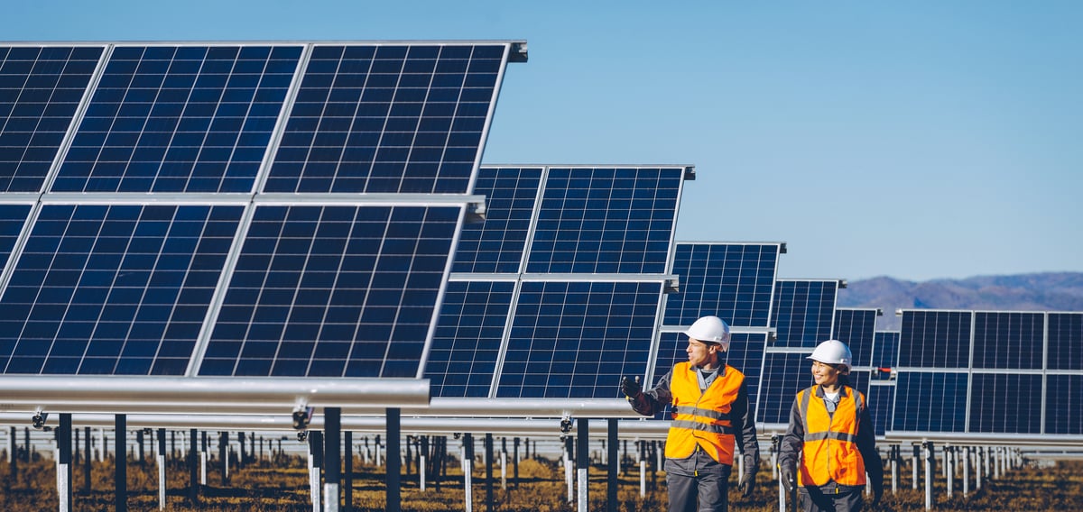 Two people wearing hard hats and orange safety vests walking beside large solar panels.