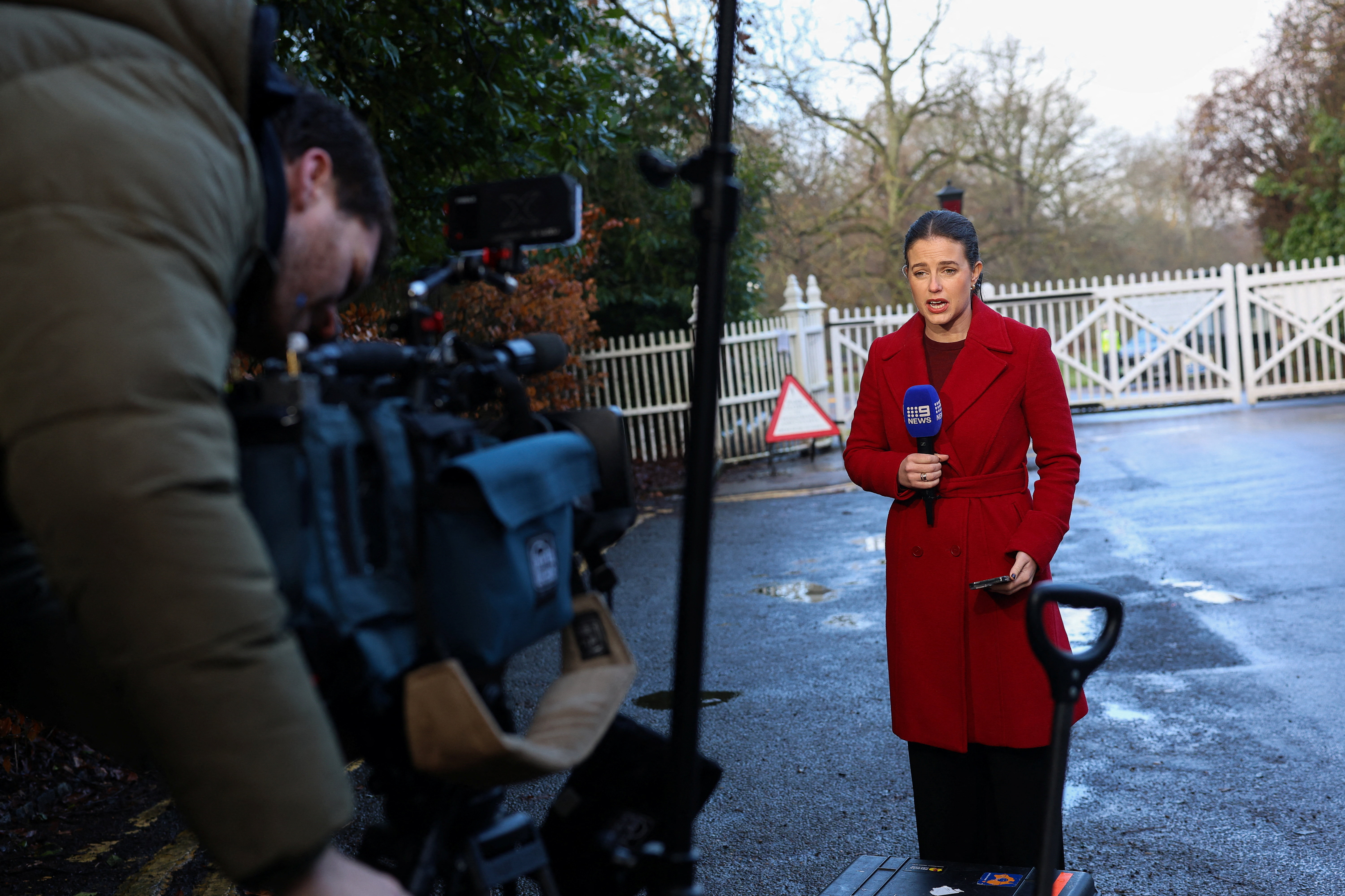 A news team reports from outside Royal Lodge in Windsor, with a female reporter holding a microphone and a cameraman in the foreground.
