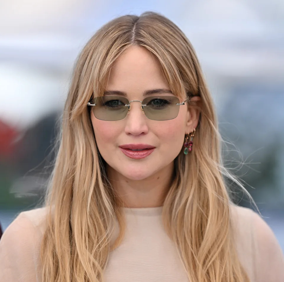 Jennifer Lawrence with long hair wearing sunglasses and earrings, dressed in a light top, attends an outdoor event
