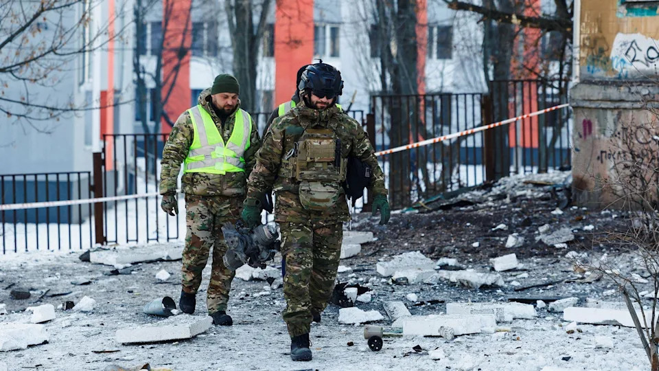 The ground is covered in snow. The police officer is wearing military clothing and there is another man behind him. the building behinf them is cordened off with tape.