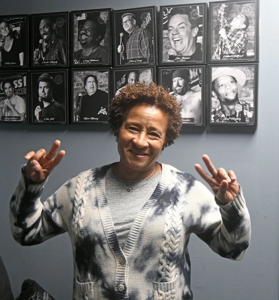 Wanda Sykes smiles and holds up peace signs, standing in front of a wall with photos of comedians performing on stage