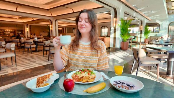An individual is seated at a table inside a restaurant, holding a cup, and smiling. The table is set with various dishes, includ An individual is seated at a table inside a restaurant, holding a cup, and smiling. The table is set with various dishes, includ