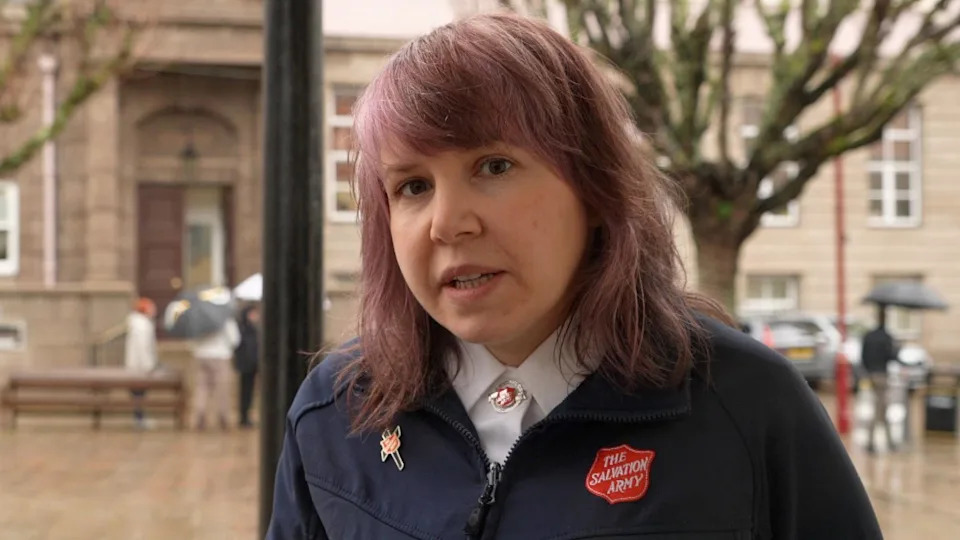 Alice Nunn is in the street. She is wearing a uniform, a navy branded coat and a white shirt with a branded brooch. She has shoulder length red hair. There are people holding umbrellas in the background.