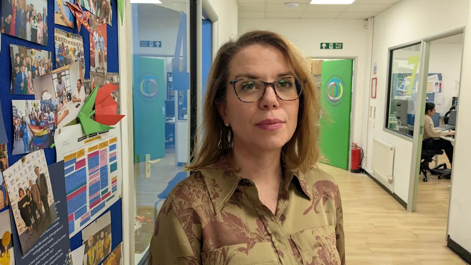 Andrea Cleaver stands in a bright corridor in the Welsh Refugee Council offices, beside a colourful bulletin board covered with photos and paper crafts. Green doors and office spaces are visible further down the hallway. She has shoulder-length light brown hair and wear glasses and pink lipstick. 