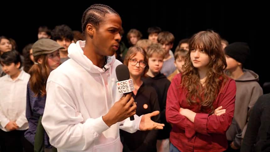 A teen journalist in a white hoodie stands holding a microphone. He is surrounded by fellow youth ready to give their answers to him. Beside him is a young person looking thoughtful with their arms folded. 