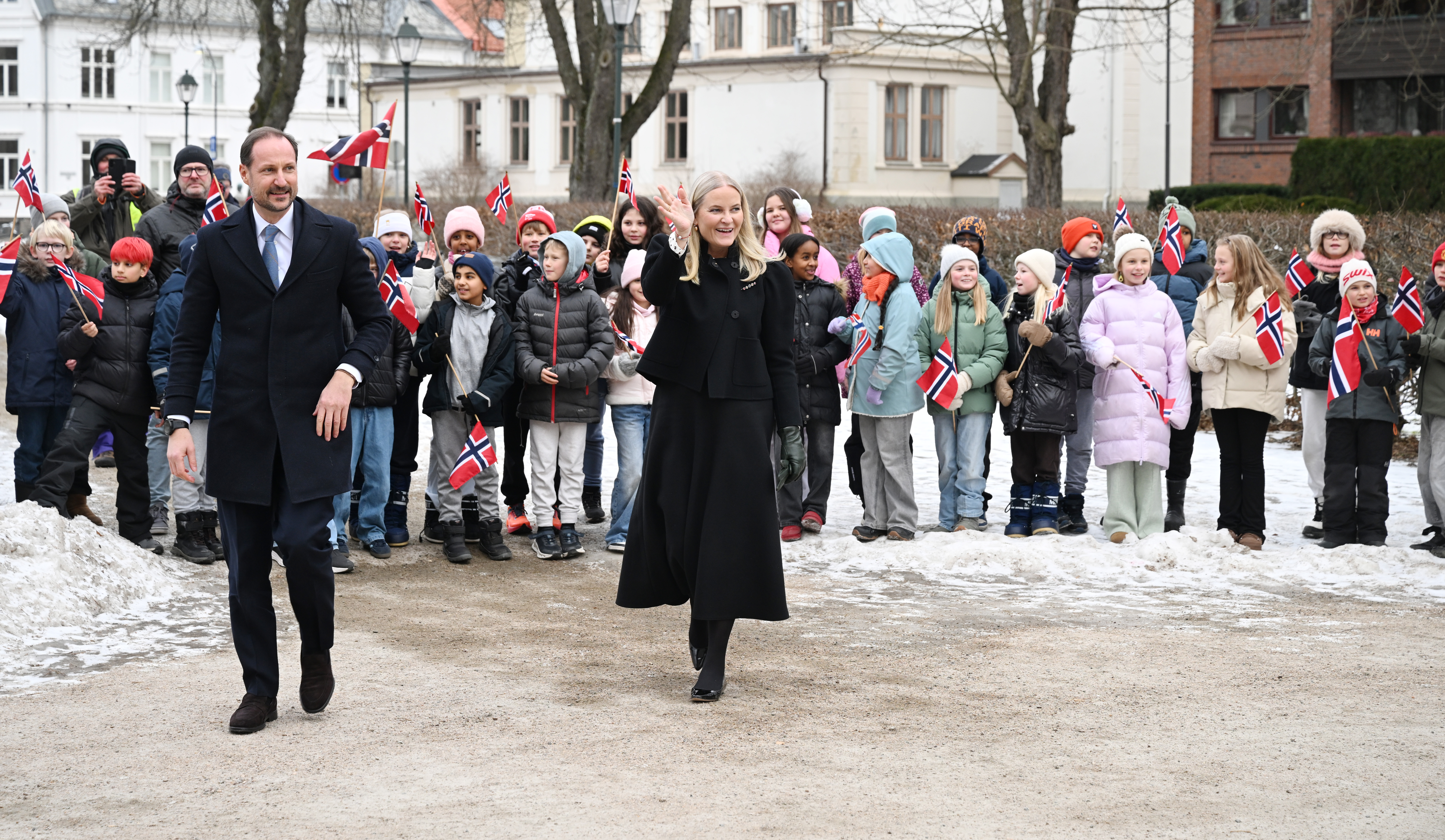 Crown Prince Haakon wearing a suit and Crown Princess Mette-Marit in a black coat waving to schoolkids outside