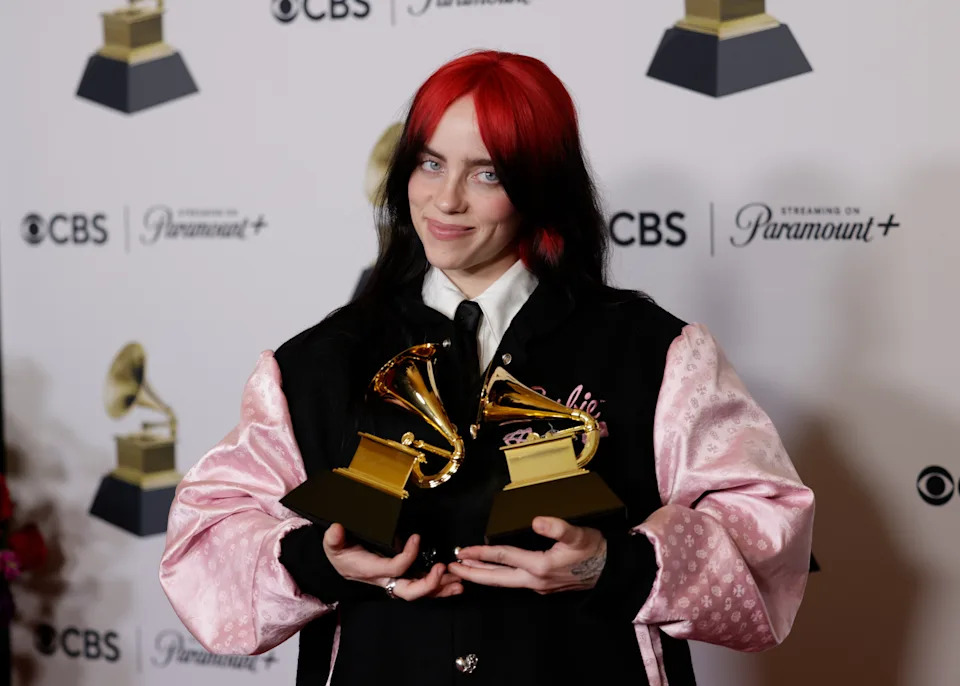 Billie Eilish holding two Grammy awards, wearing a pink and black jacket with a casual style, smiles on a press backdrop with music logos
