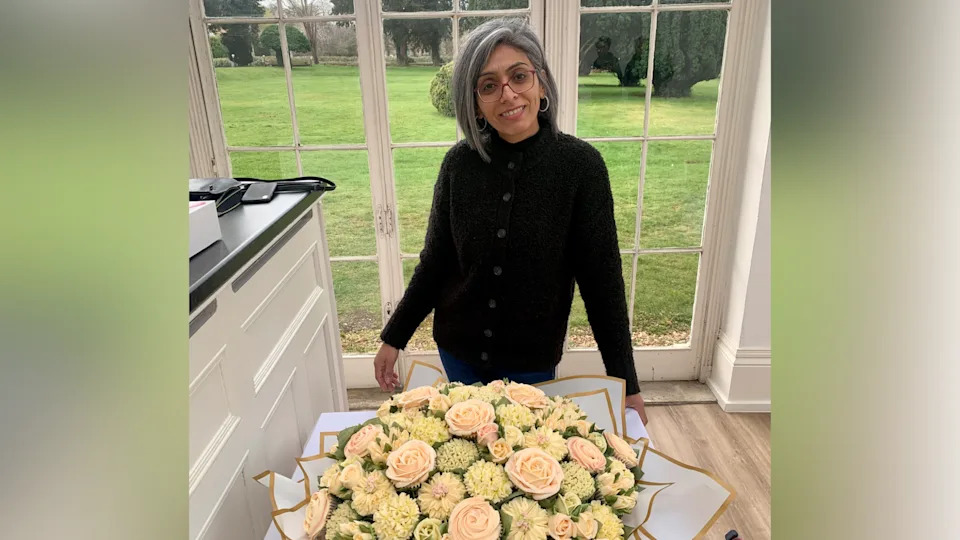 A woman with shoulder-length silver hair and glasses smiles at the camera, in a kitchen in front of glass doors. On the table in front of her is an arrangement of floral cupcakes, designed to look like a bouquet.