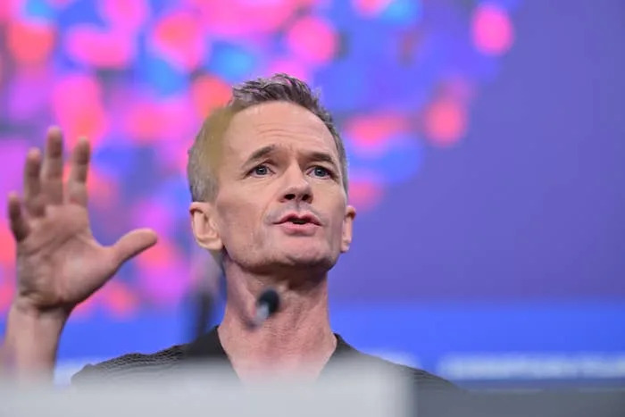 Neil Patrick Harris gestures while speaking at a conference, with abstract shapes in the background