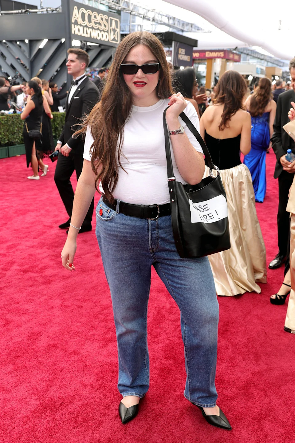 Meg Stalter on red carpet in casual outfit: white t-shirt, blue jeans, black heels, sunglasses, and tote bag