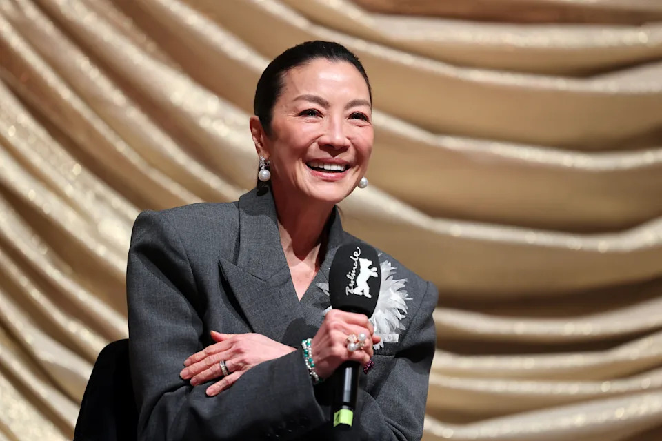 Michelle Yeoh smiling while speaking into a microphone, wearing a tailored blazer with feather detail, sitting in front of draped fabric background