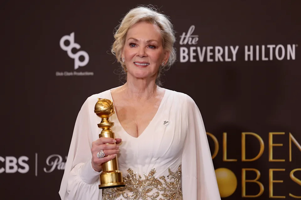 Jean Smart in an elegant gown holding an award at a formal event, posing in front of a backdrop with event sponsors