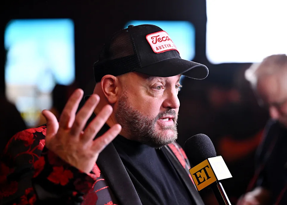 Kevin James wearing a patterned jacket and cap, speaking into an ET microphone at an event