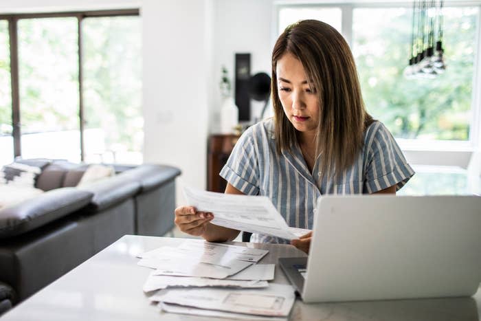A woman reviews financial documents at home, sitting by a laptop on a table with scattered papers