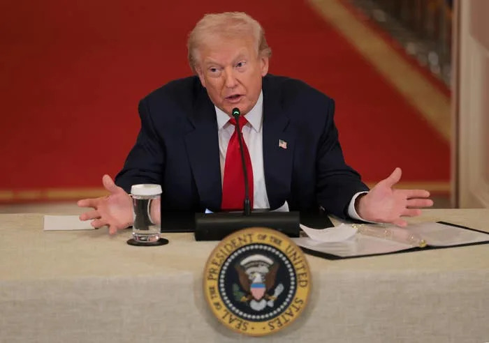 Donald Trump speaking at a table with a microphone and glass of water, seated in front of a lectern with a presidential seal