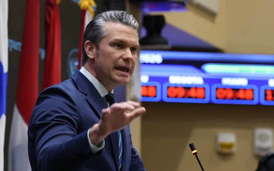 A man in a suit gestures while speaking at a podium in a formal setting. Digital displays are visible in the background
