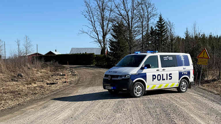 Finnish police van blocking a country road, with houses, trees and blue sky seen in the background.