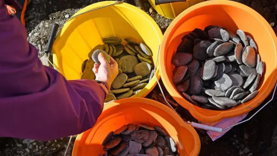 Shows several bright buckets filled with smooth, flat stones, with a person selecting one from a yellow bucket.