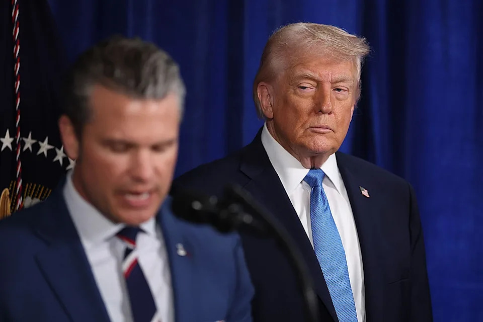 Man in suit speaks at podium; another man in suit stands behind, looking on attentively