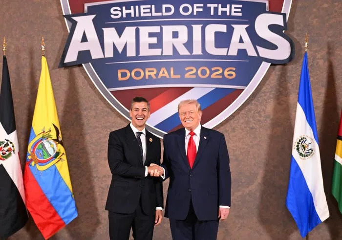 Donald Trump shaking hands at a diplomatic event, standing before flags from various countries, with a "Shield of the Americas Doral 2026" sign