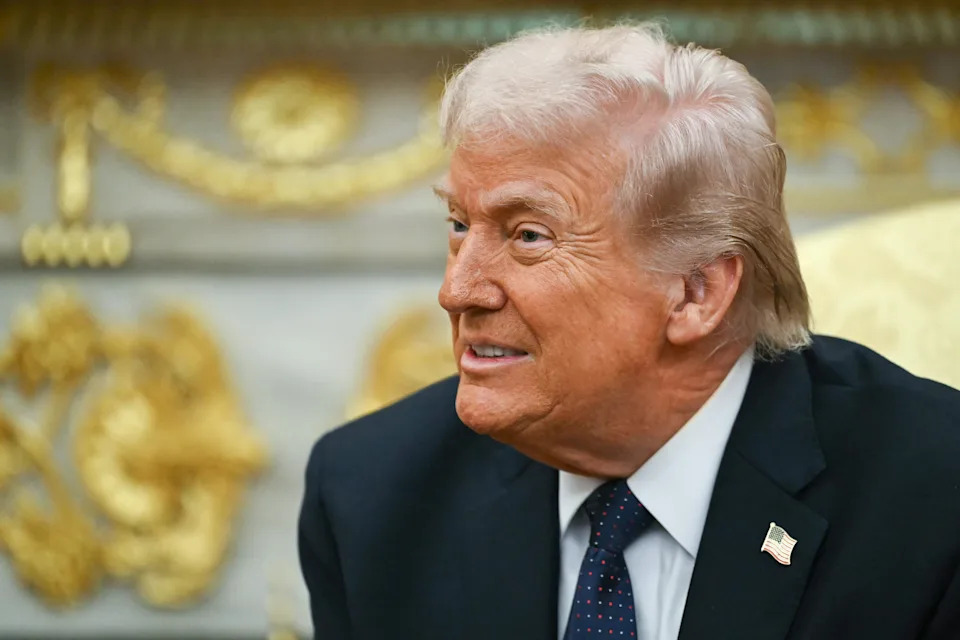Donald Trump in a suit with an American flag pin is sitting and smiling, appearing engaged in conversation