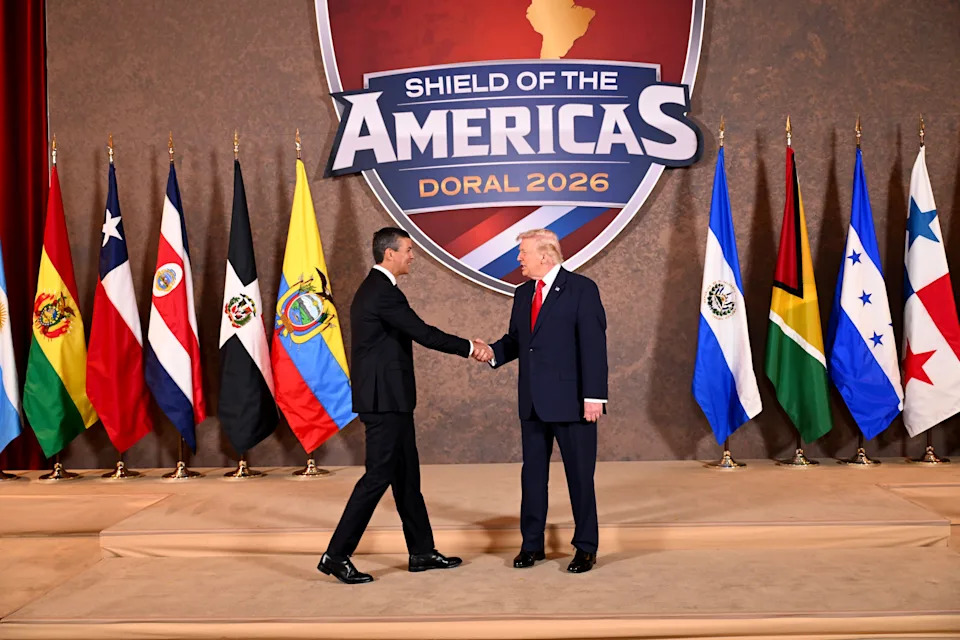 Santiago Pena and Donald Trump shake hands on a stage labeled "Shield of the Americas Doral 2026," with multiple flags displayed in the background