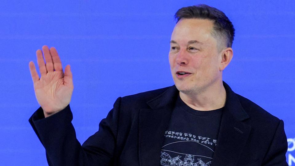 Elon Musk waving to a crowd from a stage wearing a black blazer and a black t-shirt.