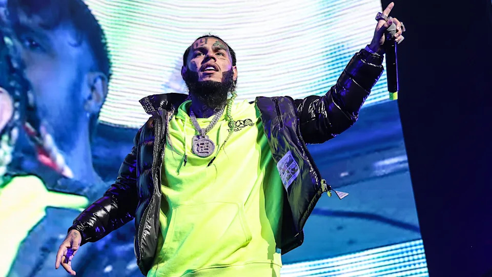 6ix9ine on stage wearing a neon green hoodie and black jacket, holding a microphone with a large chain necklace.