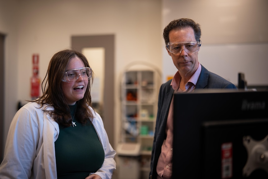 Eric May in the lab, looks seriously at a computer, alongside a smiling woman, both wear protective glasses.
