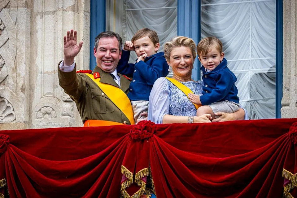 Grand Duke Guillaume waving while standing with Grand Duchess Stephanie on a balcony. The pair are holding their young children
