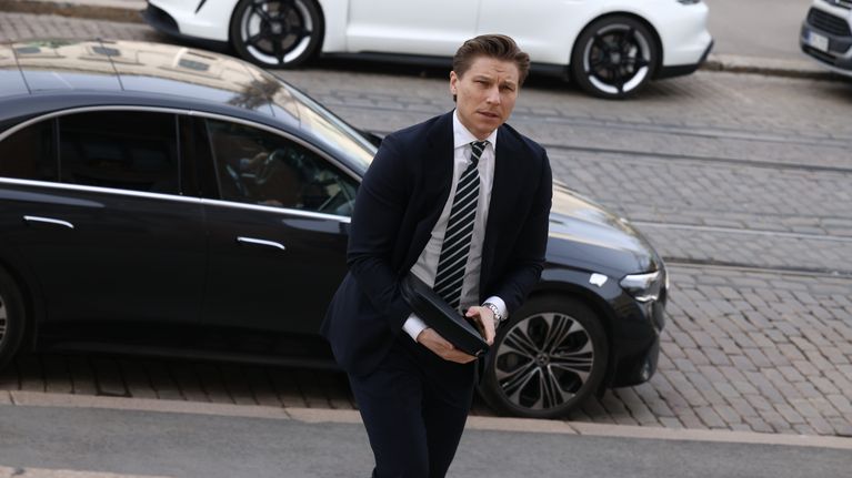 Man in dark suit, white shirt and striped necktie walking up steps, with two cars and cobblestone road in the background.