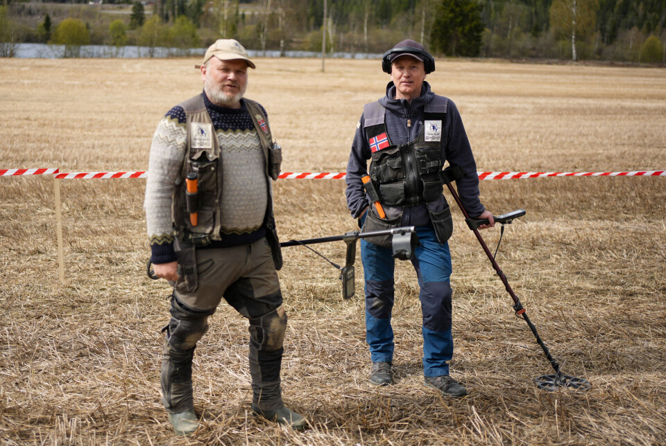 Two detectorists stand in a field with metal detectors and a red-and-white tape barrier behind them.