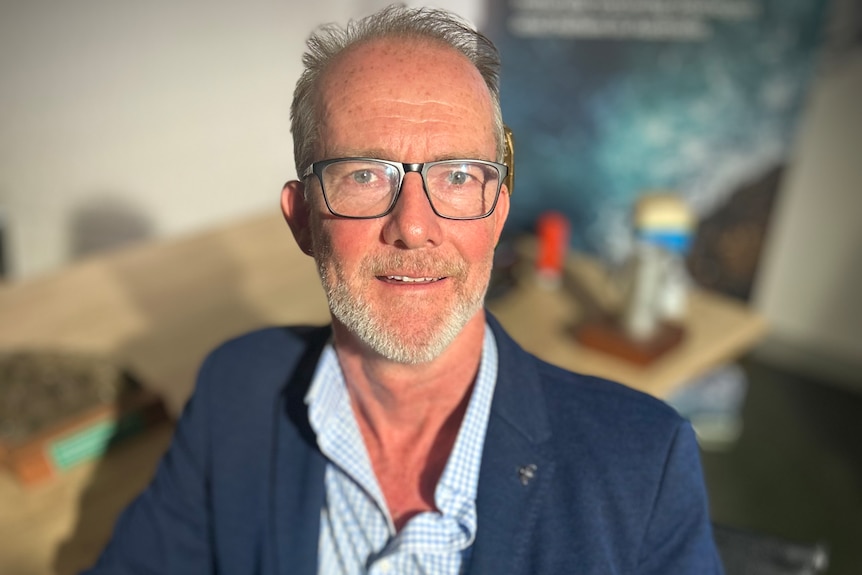 Neil smiles, wears blue jacket over light blue shirt, balding, grey beard, glasses, Anglo-Saxon, blurred desk in background.