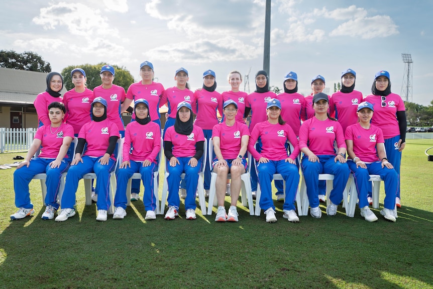 Two rows standing and seated of Afghan women cricket atheletes in hot-pink and blue uniform, on green sports field.