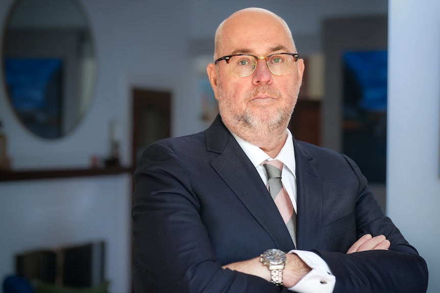 A man wearing a navy suit and glasses stands with his arms crossed in front of his chest.