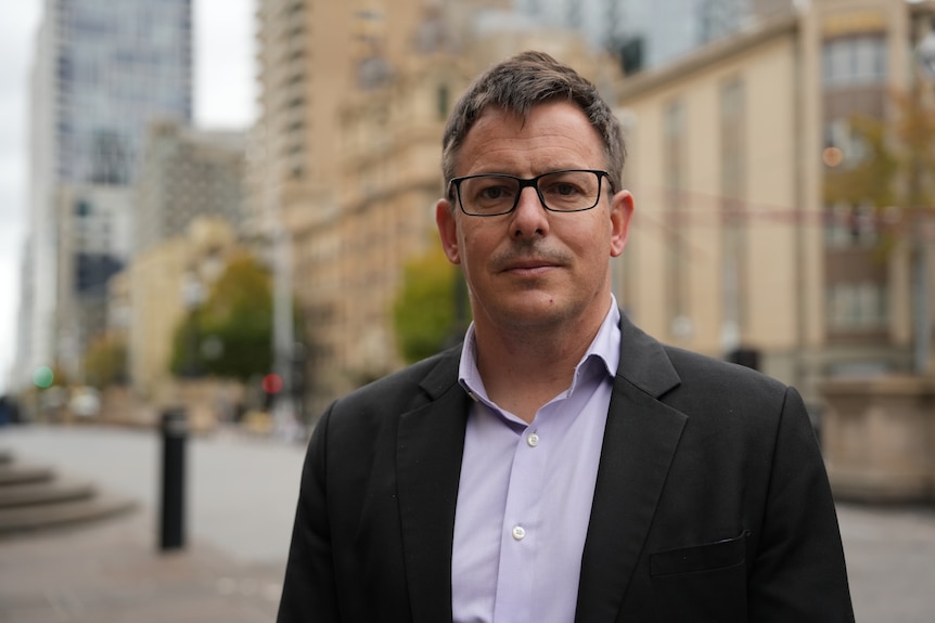 A man with short dark hair and glasses stands outdoors looking serious.